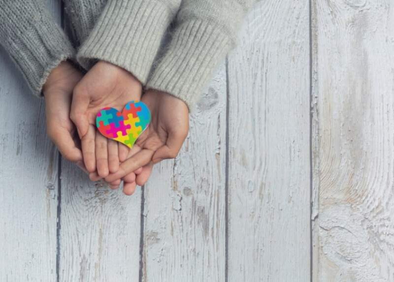 Two hands holding a heart-shaped puzzle piece symbolizing autism, made of colorful pieces.