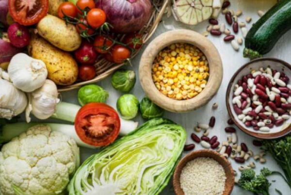 A variety of fresh vegetables, legumes, and grains, including cabbage, beans, and tomatoes, displayed on a wooden surface.