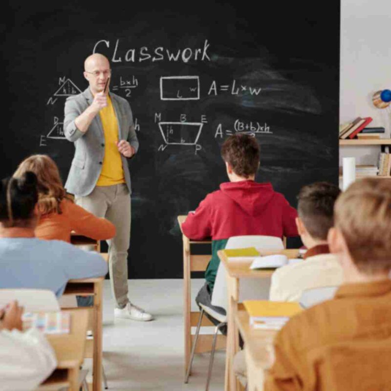 A classroom with a teacher teaching on a blackboard while students listen attentively, raising their hands to ask questions.