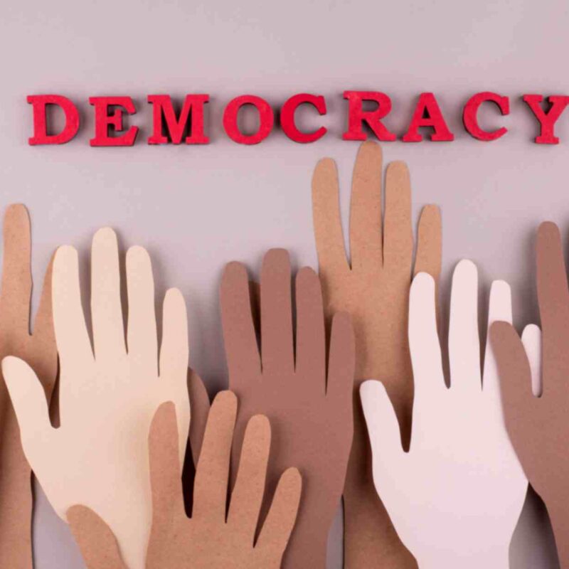 Diverse paper hands raised beneath the word "DEMOCRACY," symbolizing inclusivity, civic engagement, and democratic participation.