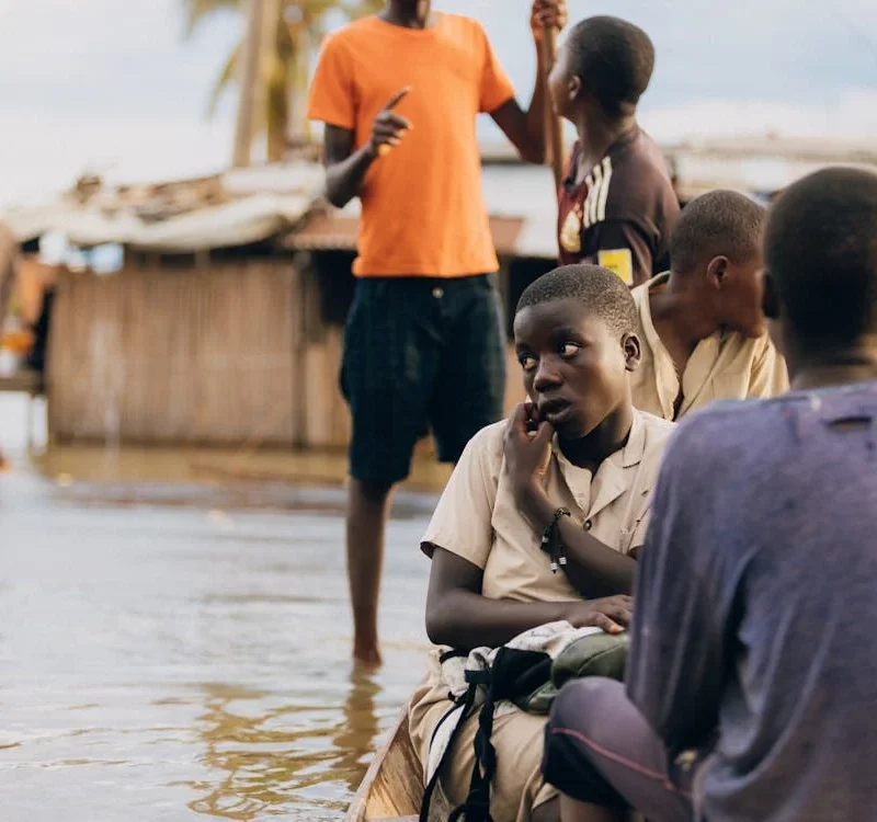 Children sit in shallow floodwater near damaged structures, highlighting the effects of climate change on vulnerable communities.