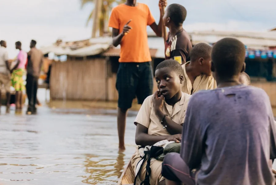 Children sit in shallow floodwater near damaged structures, highlighting the effects of climate change on vulnerable communities.