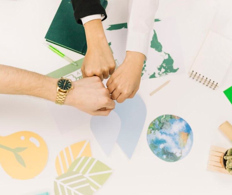 Three hands fist-bumping over a desk with environmental symbols, representing teamwork in global climate action projects.