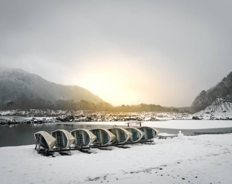 Snow-covered boats by a tranquil lake with mountains and a golden sunrise, symbolizing resilience in the Himalayan region.