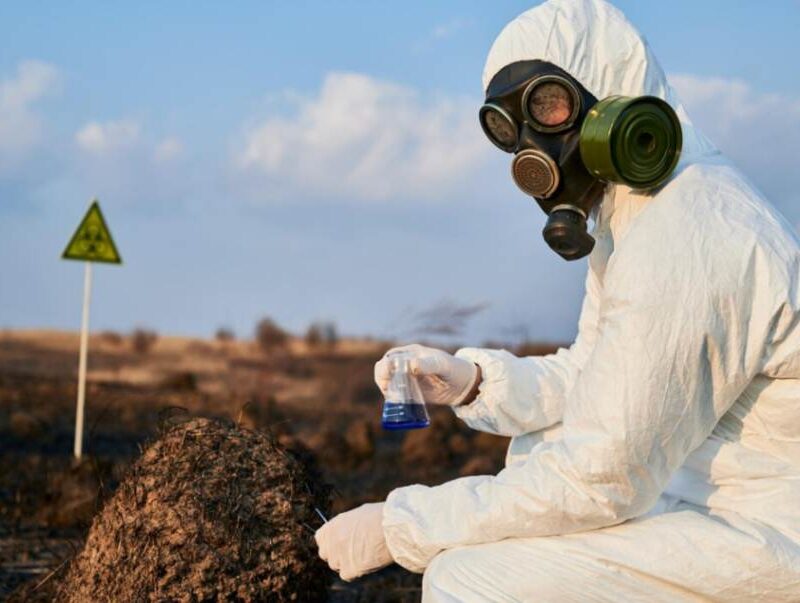 Person in protective suit and gas mask holding a test flask in a burnt field, symbolizing chemical threat detection and analysis.