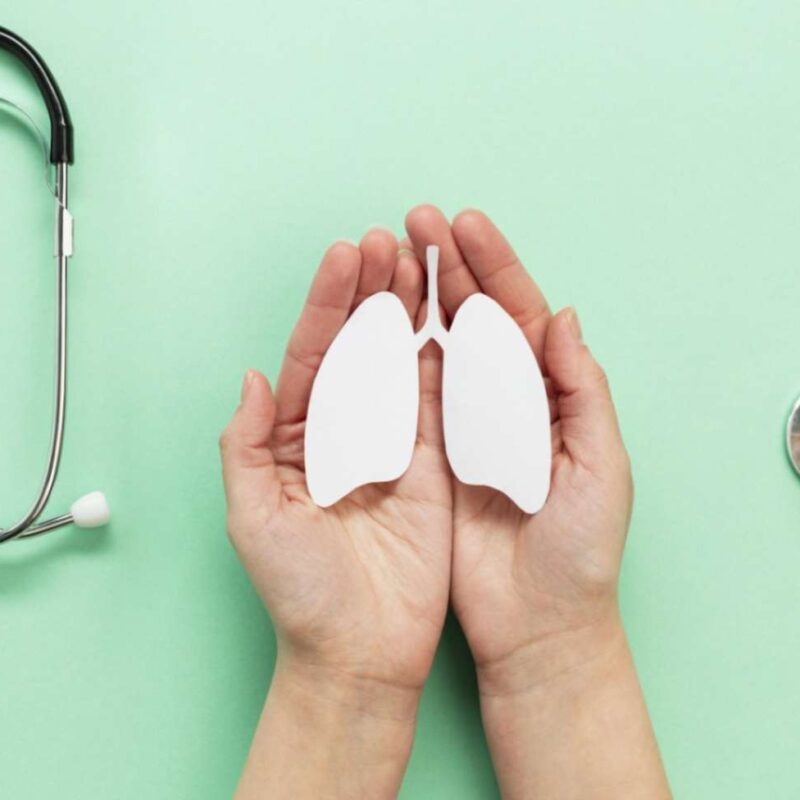 Hands holding a white cutout of lungs on a light green background, accompanied by a stethoscope symbolizing healthcare and TB awareness.